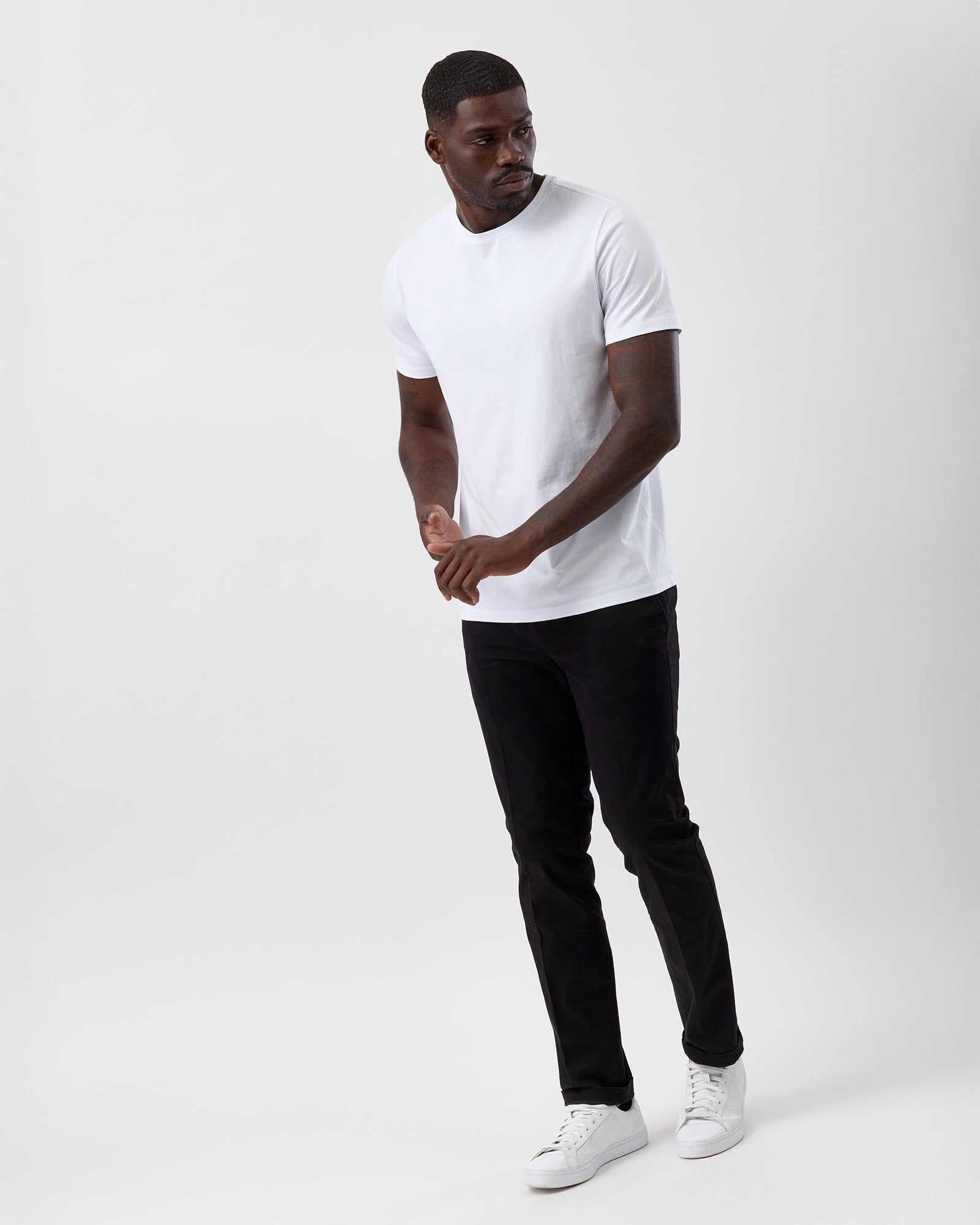 Man wearing a white t-shirt, black cotton stretch chino pants, and white sneakers on a plain background, street styled, full body shot