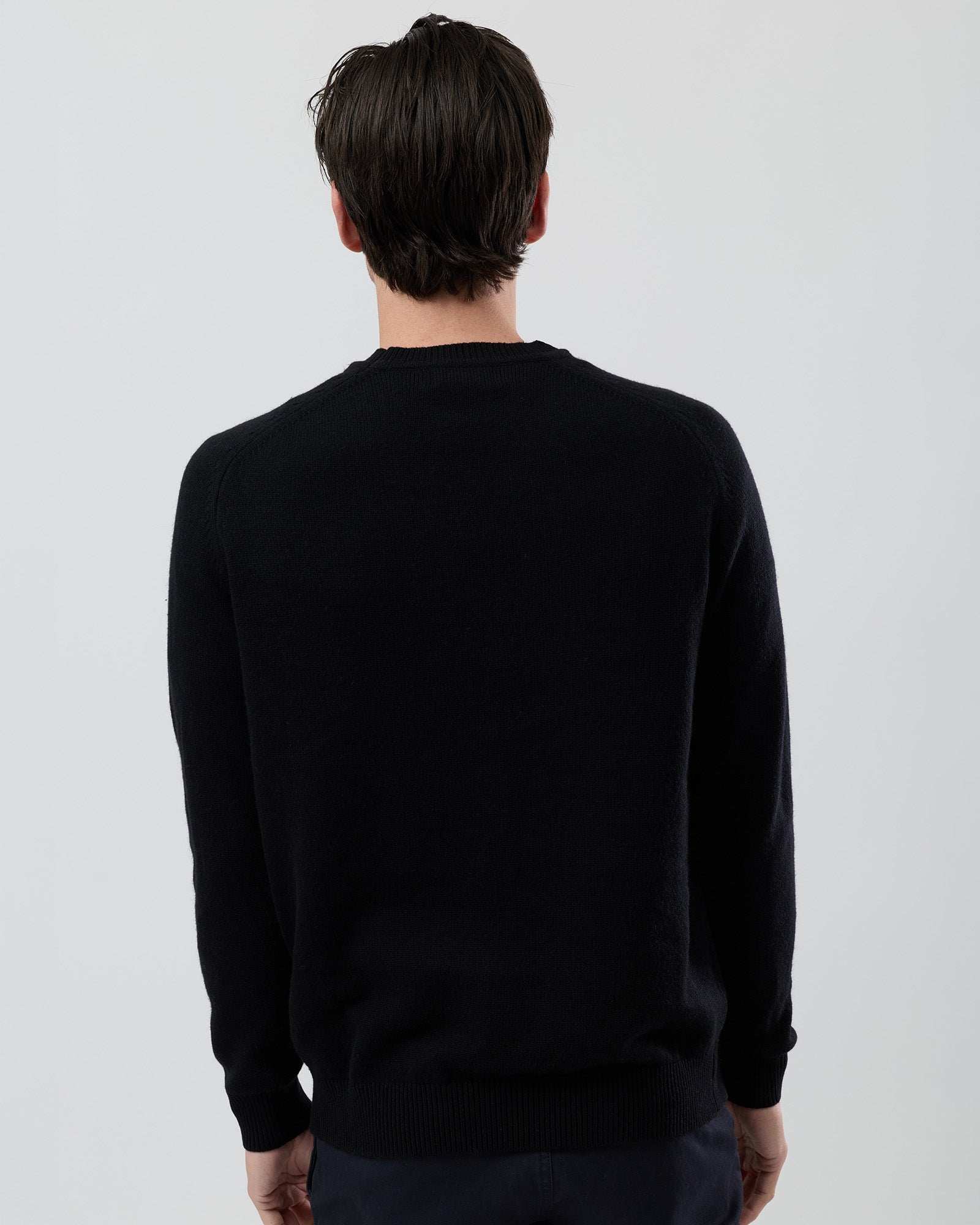 Rear view of a men's black wool blend crew neck sweater highlighting the saddle shoulder construction and clean fit on a grey background.