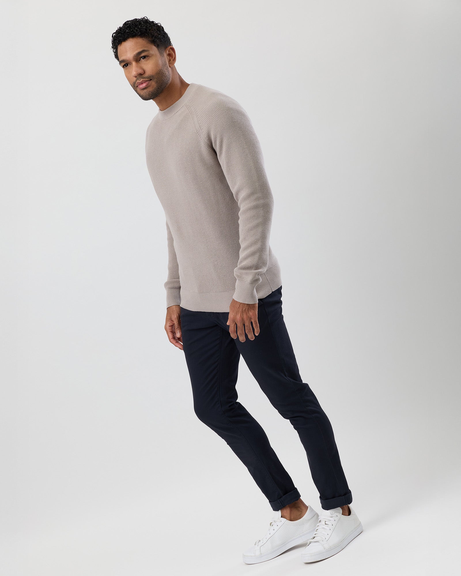 Man wearing a beige sweater, black pants, and white sneakers on a white background