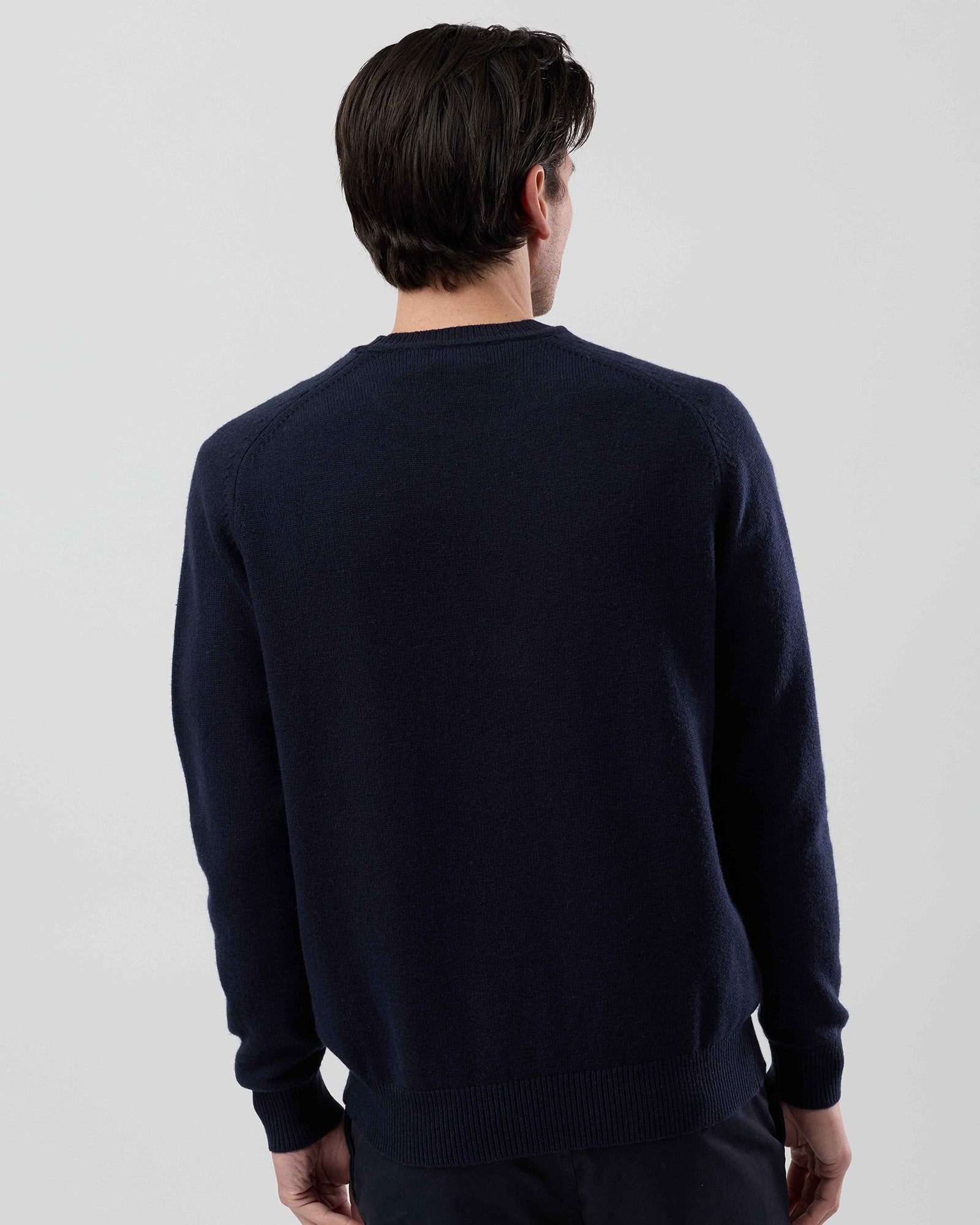 Rear view of a men's navy wool blend crew neck sweater highlighting the clean silhouette and saddle shoulder construction on a grey background.