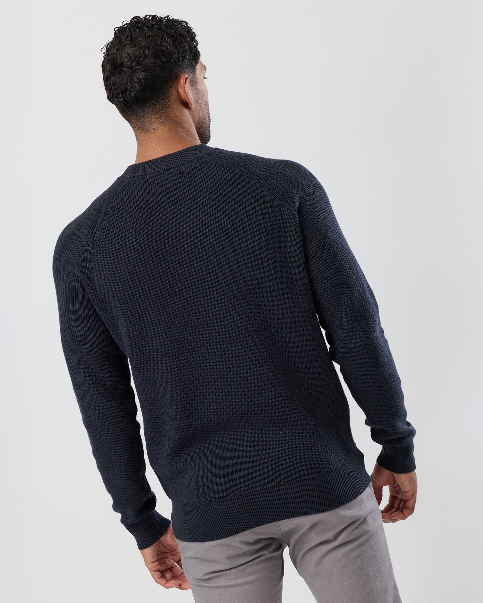 Man wearing a navy fisherman's rib sweater and light gray pants on a white background, rear view