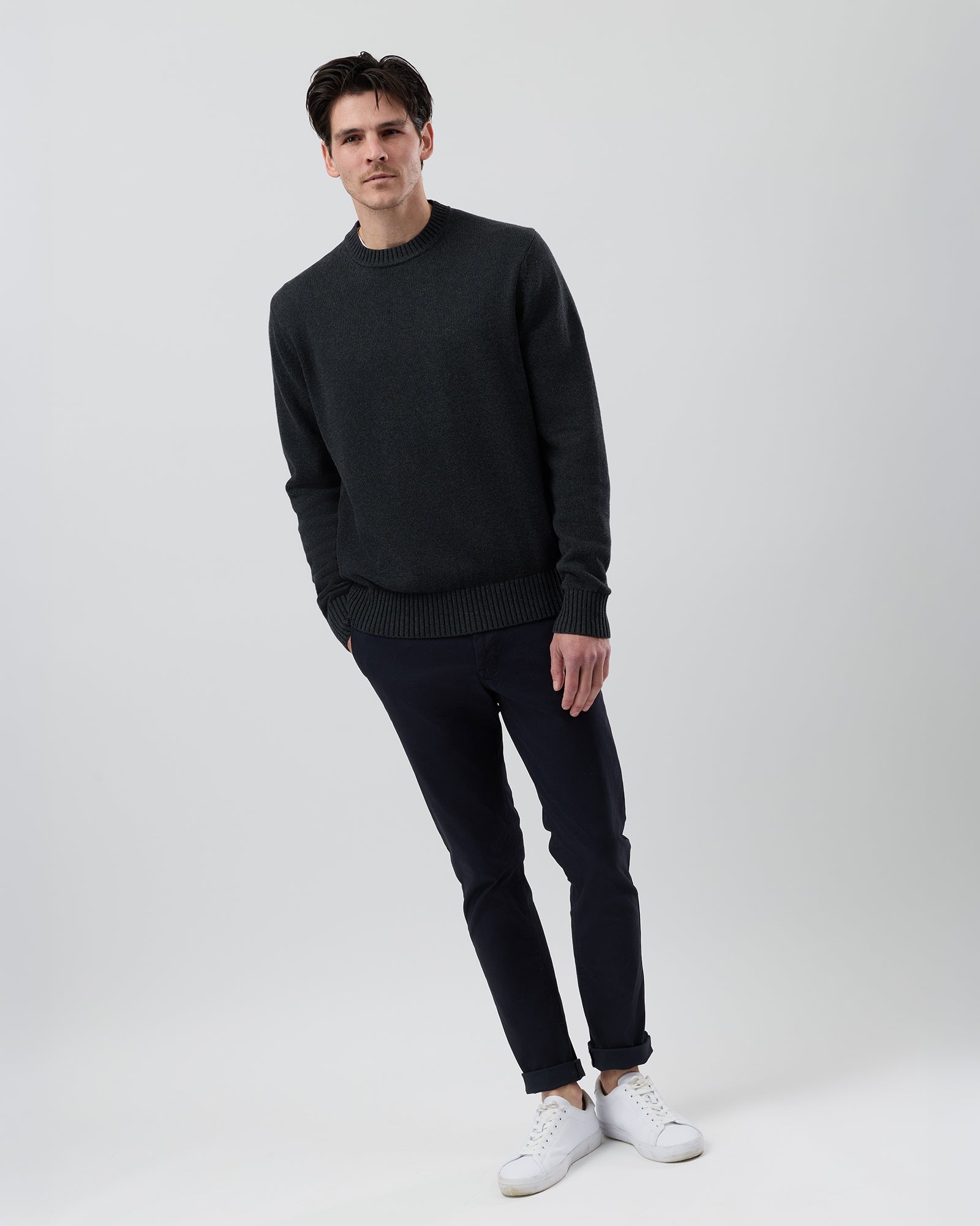 Full body view of a male model wearing the Wayver charcoal Essential Cotton Crew Neck Knit styled with navy chinos on a grey background.