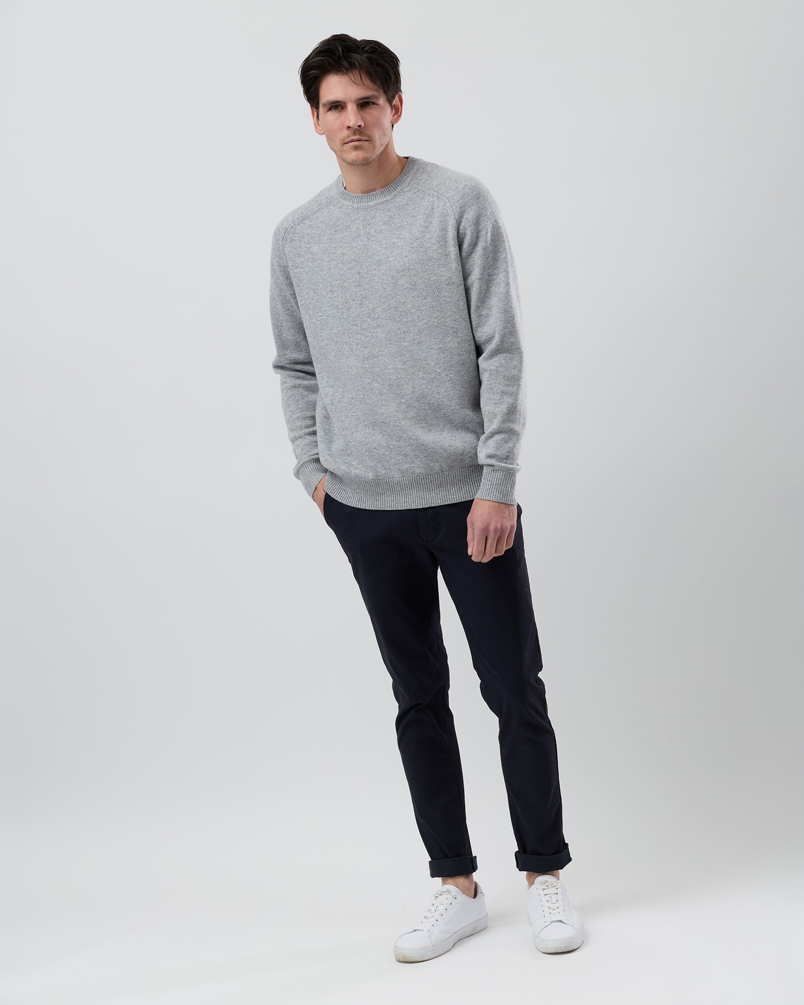 Full-length front view of a male model wearing the Wayver light grey wool blend crew neck knit styled with navy chinos on a grey background.