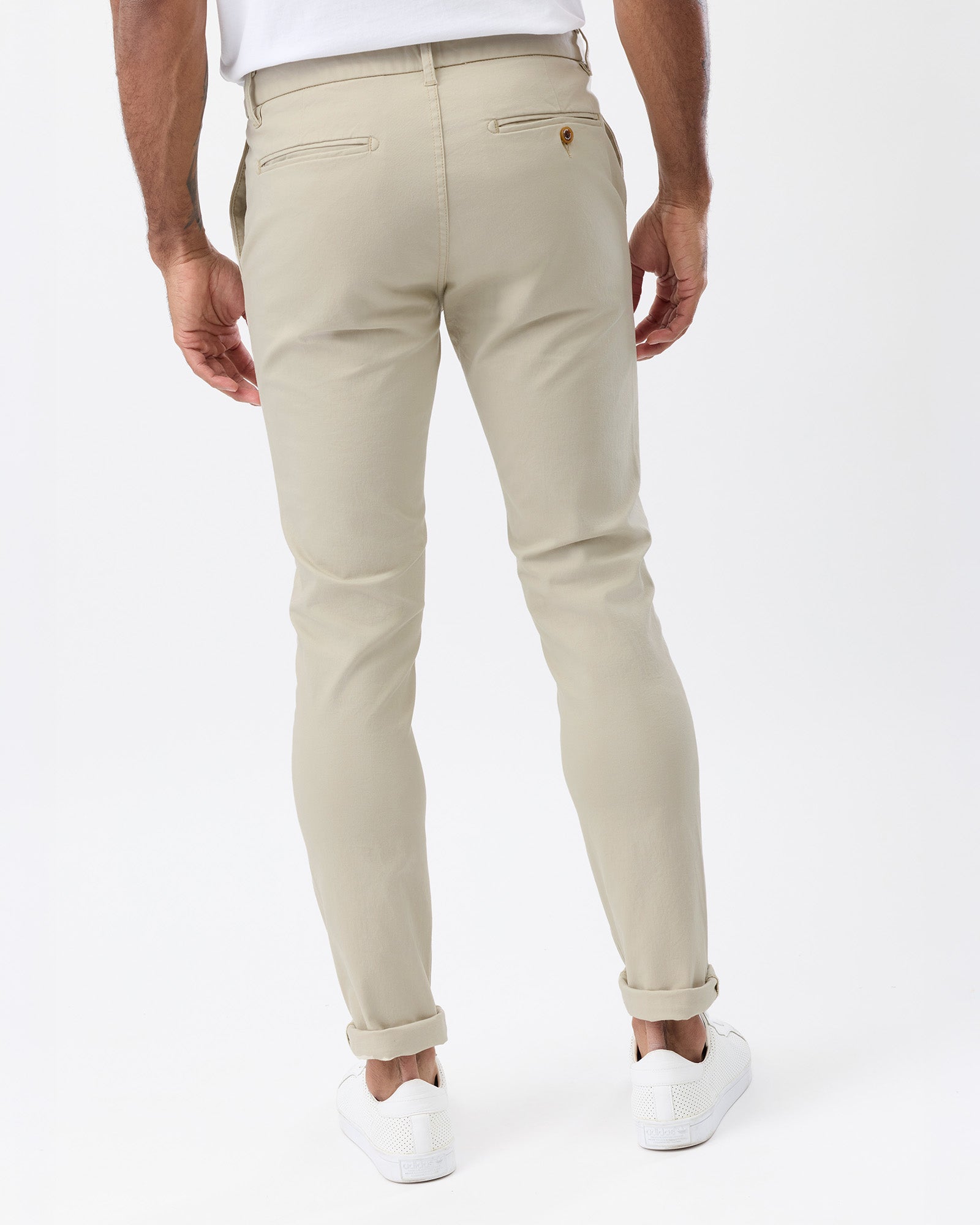A man wearing beige slim-fit chino pants, shown from the back. The pants feature a classic five-pocket design, belt loops, and rolled cuffs, paired with a white t-shirt and white sneakers against a clean white studio background.