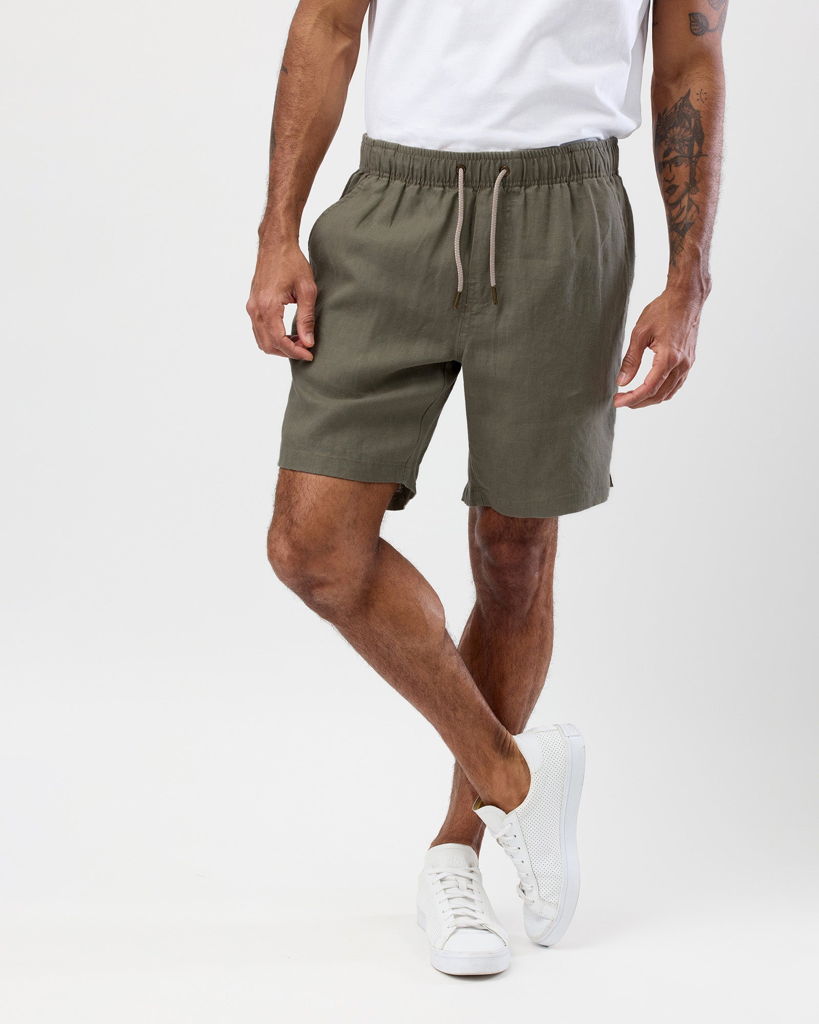 A man wearing olive green drawstring shorts, a white t-shirt, and white sneakers, standing against a plain white studio background.