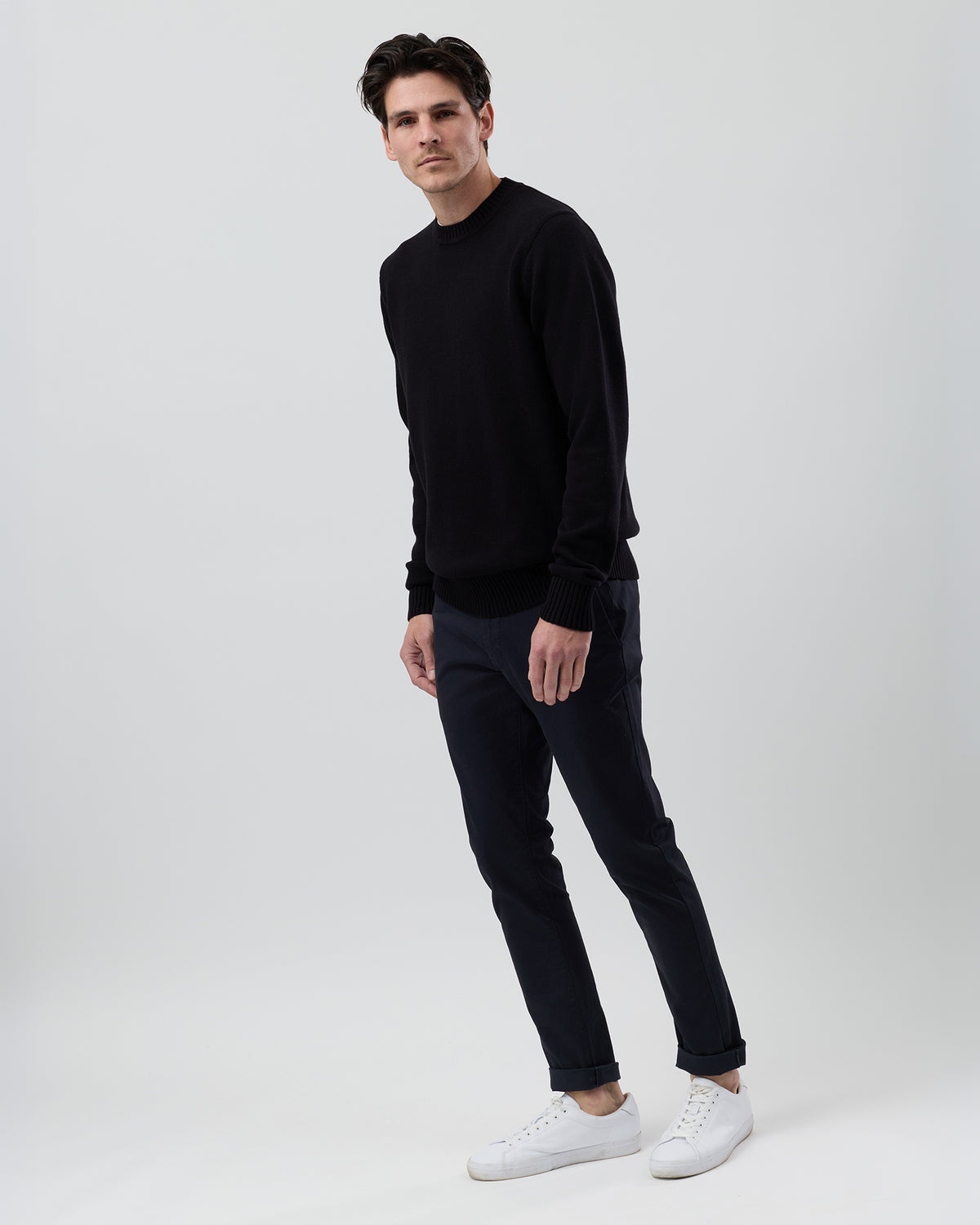 Full-length side profile of a men's black cotton crew neck jumper paired with navy chinos on a grey background.
