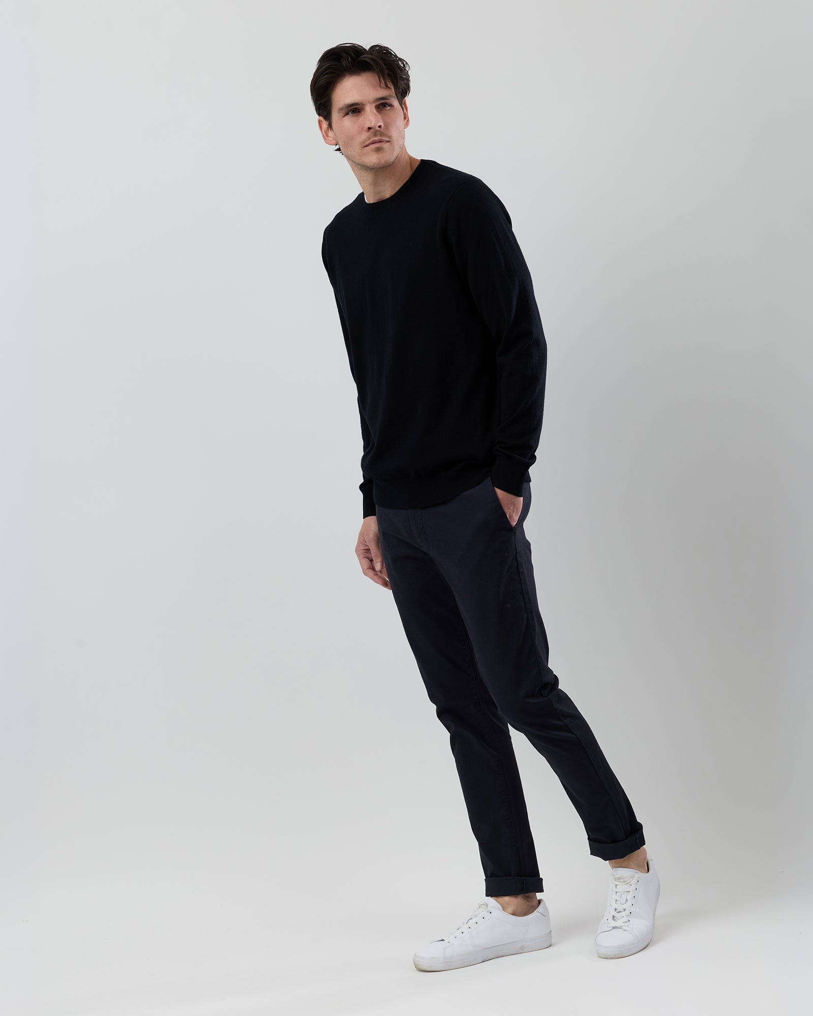Full-length side profile of a men's black Australian ultrafine merino wool crew neck knit styled with navy chinos on a grey background.
