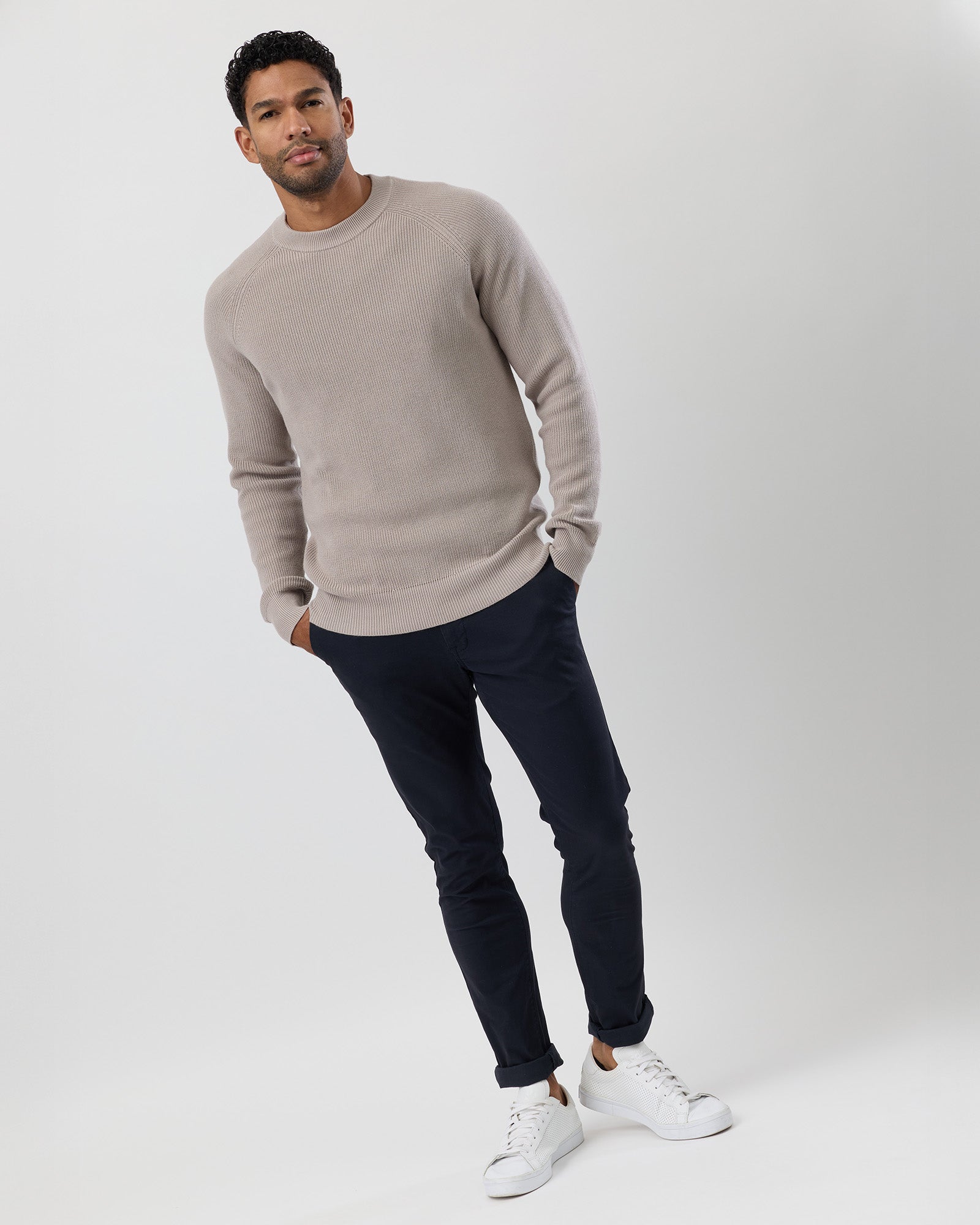 Man wearing a beige sweater, dark chinos, and white sneakers on a white background