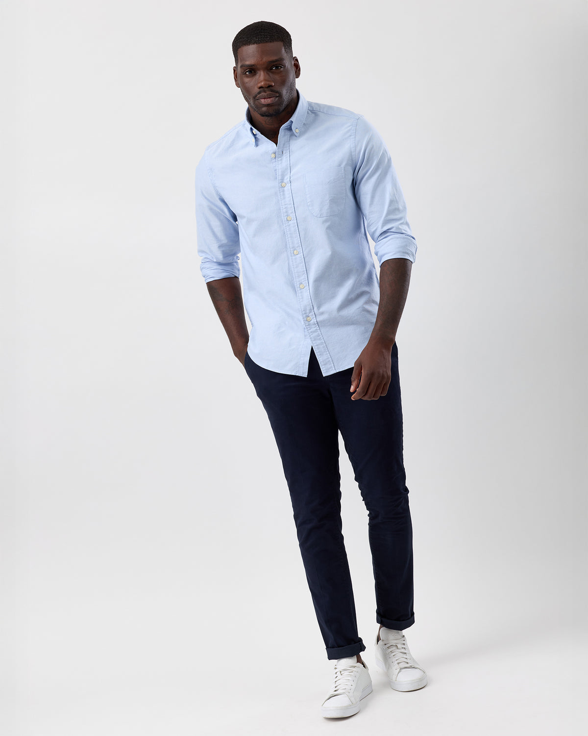 Full length image of a man wearing a light blue cotton oxford shirt with sleeves rolled, navy chinos and white sneakers.