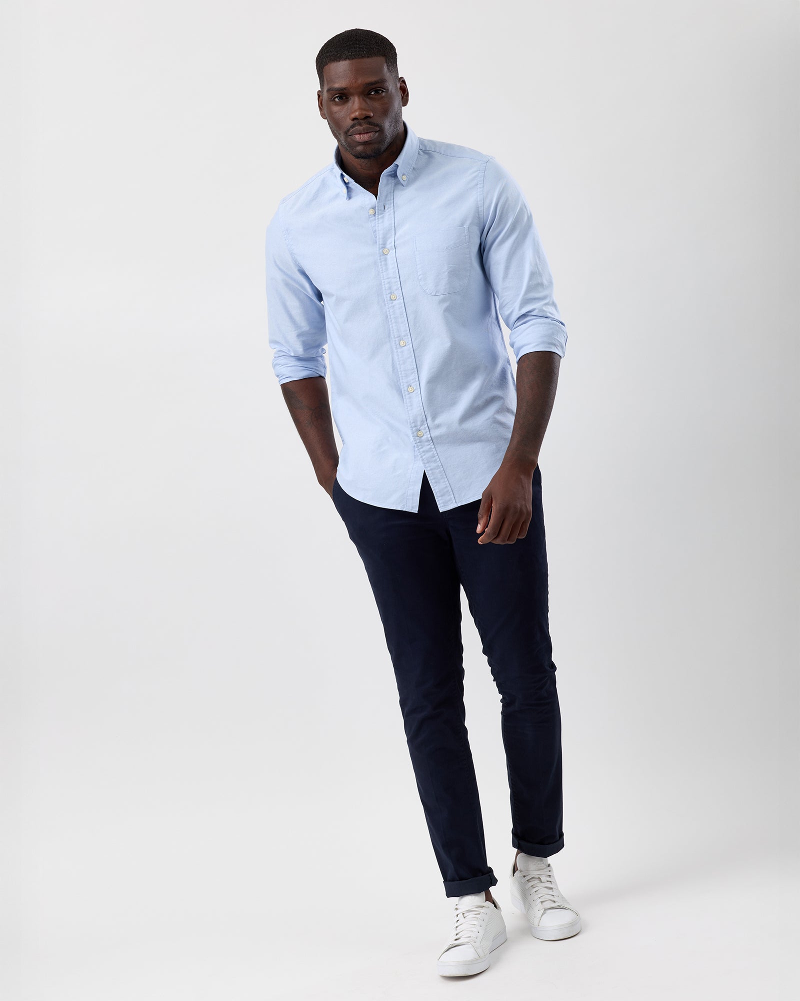 Full length image of a man wearing a light blue cotton oxford shirt with sleeves rolled, navy chinos and white sneakers.