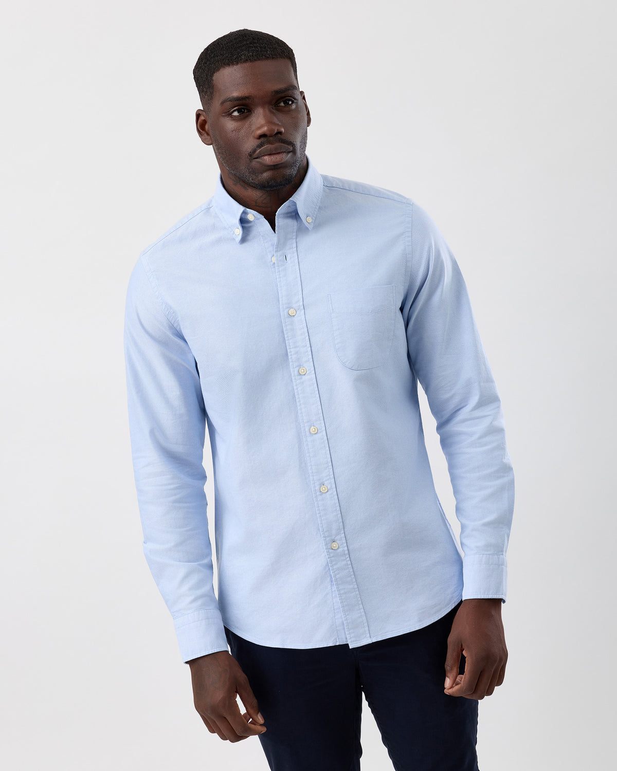 Man wearing a light blue button-up cotton oxfrod shirt against with navy chinos a plain background