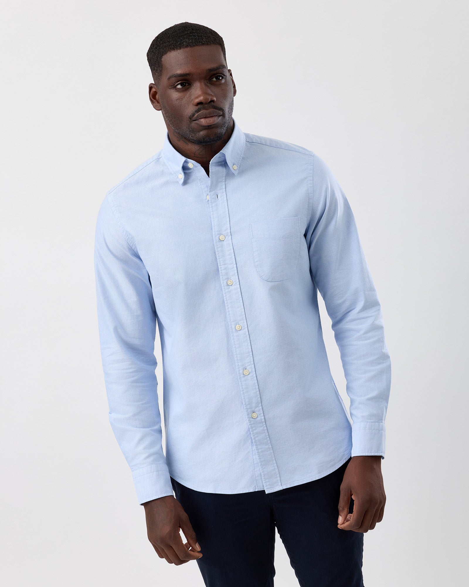 Man wearing a light blue button-up cotton oxfrod shirt against with navy chinos a plain background