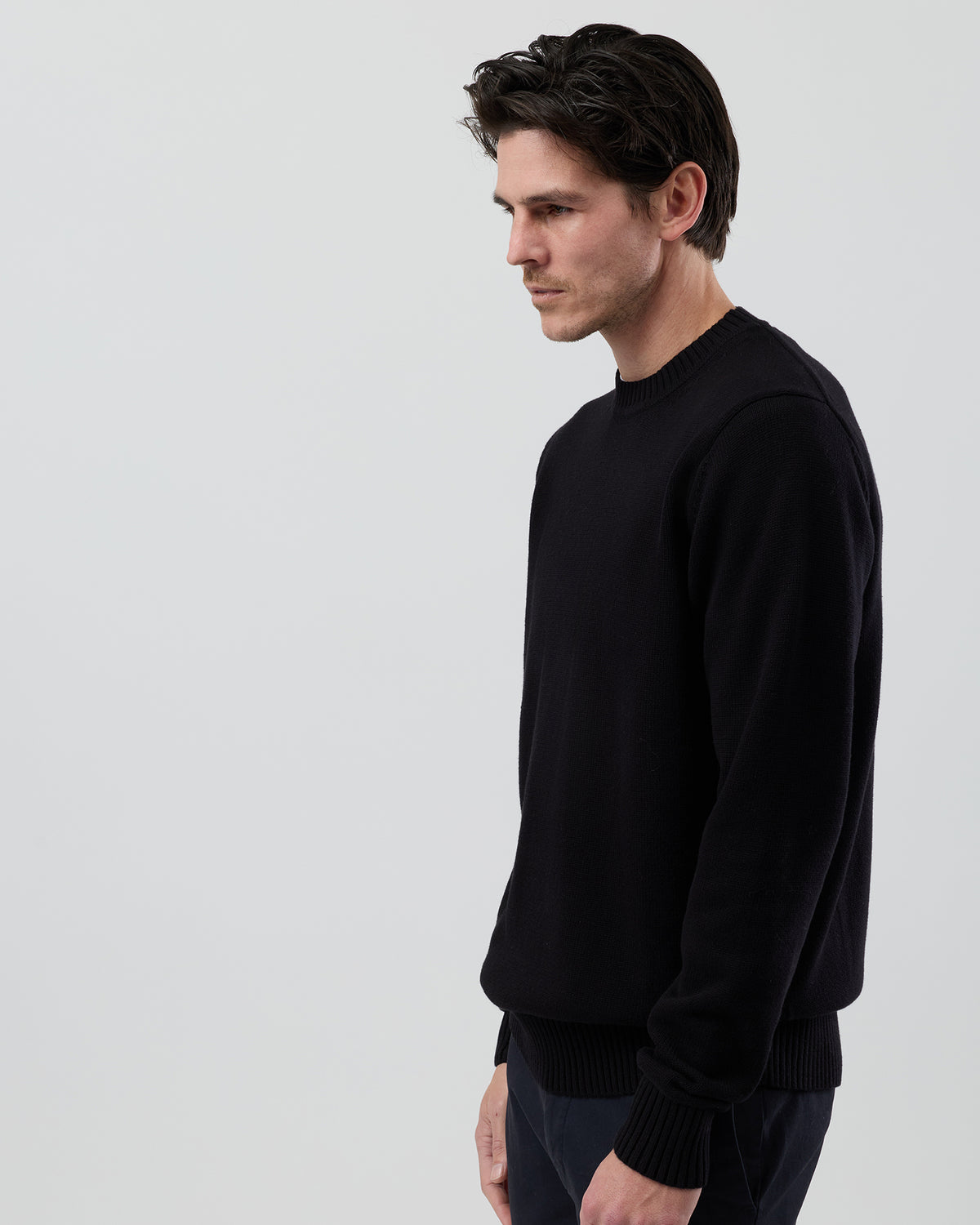 Angled side view of a men's black cotton crew neck knit showing the clean silhouette and tailored sleeve fit against a grey background.