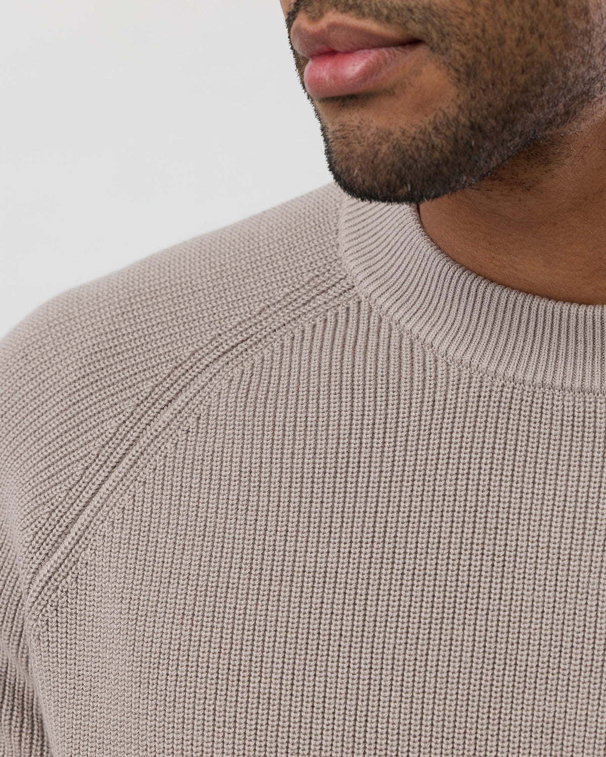 Close-up of a person wearing a beige knitted sweater against a light gray background - showing stitch detail