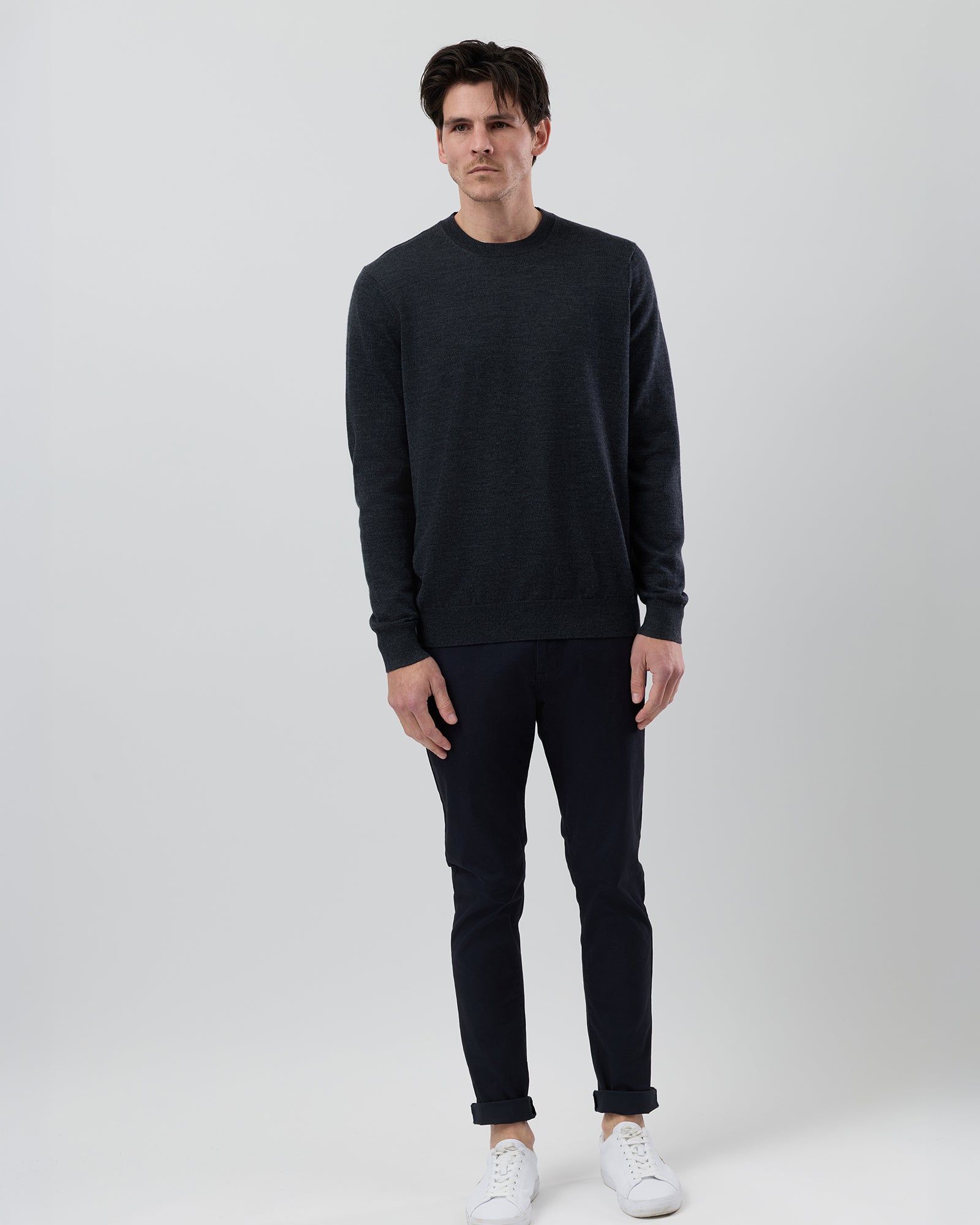 Full-length front view of a male model wearing a dark charcoal marl Australian ultrafine merino crew neck knit styled with navy chinos on a grey background.