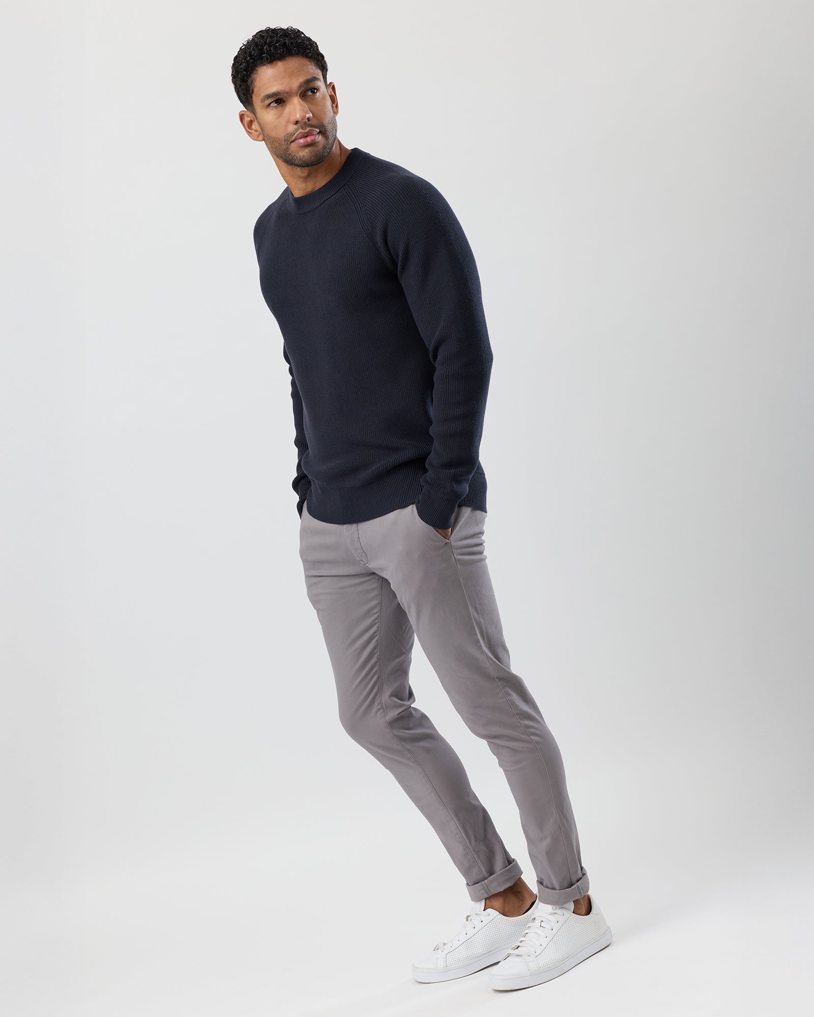 Male model wearing fisherman's crew knit in dark navy, with grey chinos and white sneakers, full body view