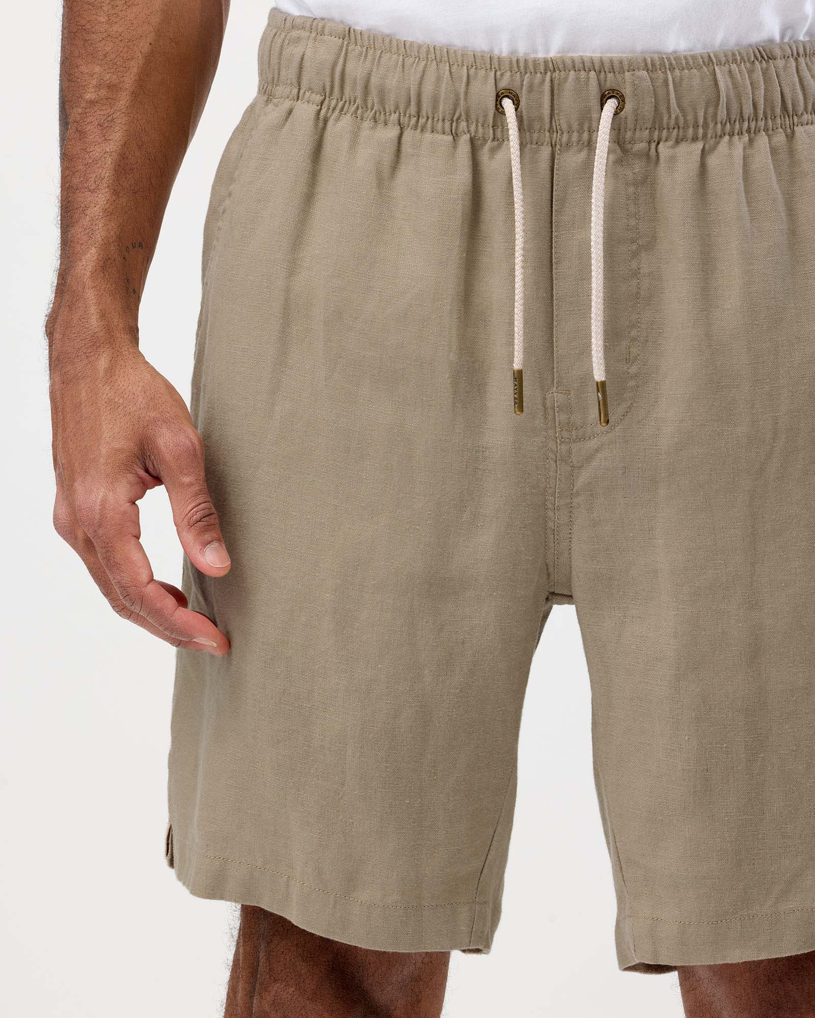 Close-up of a person wearing light khaki drawstring shorts, showing the waistband, fabric texture, and part of the hand and legs against a plain light background.