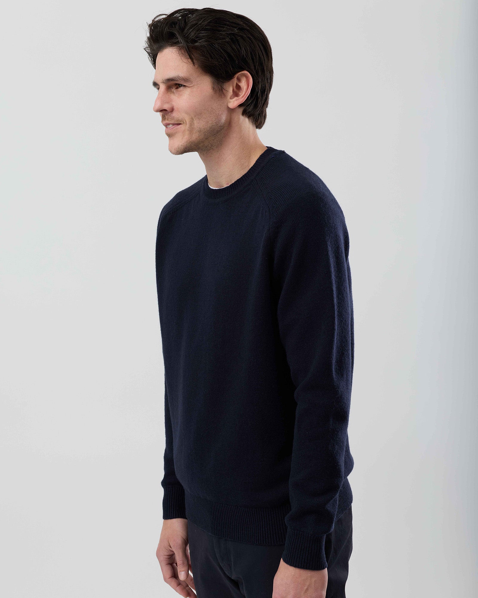 Angled side view of a men's navy wool blend crew neck knit showing the tailored fit and premium knit construction against a grey background.