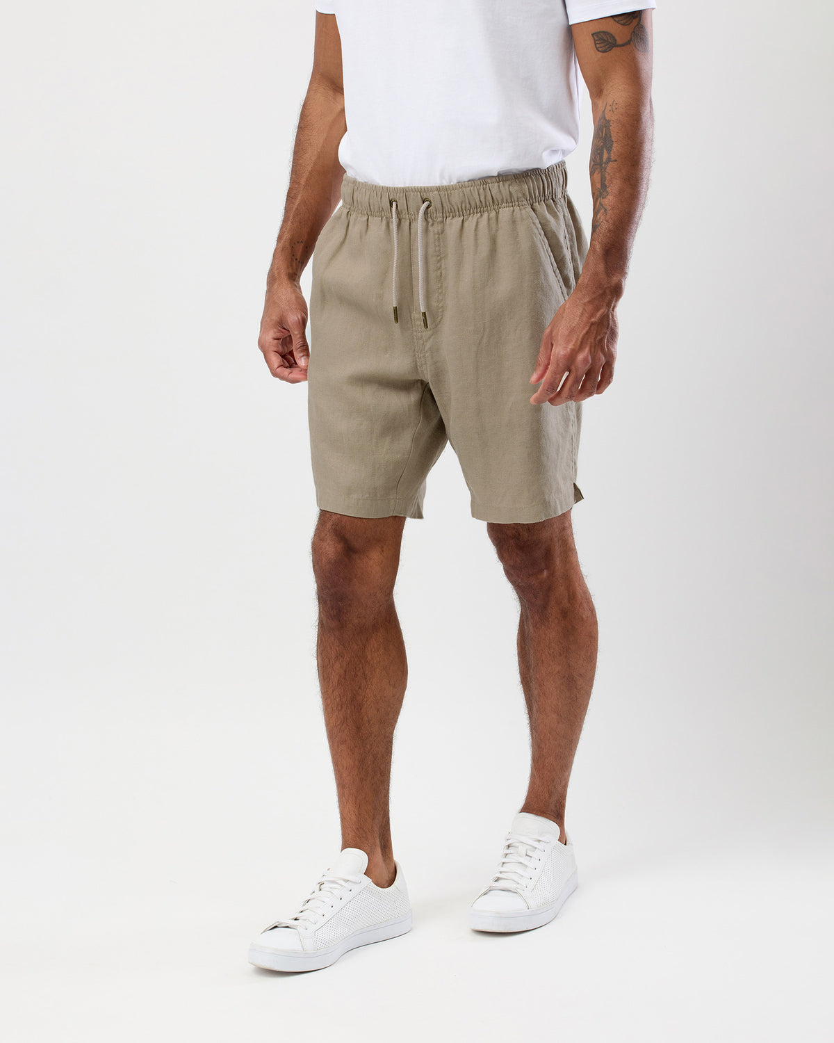 A man wearing beige drawstring shorts, a white t-shirt, and white sneakers, standing against a light gray studio background.