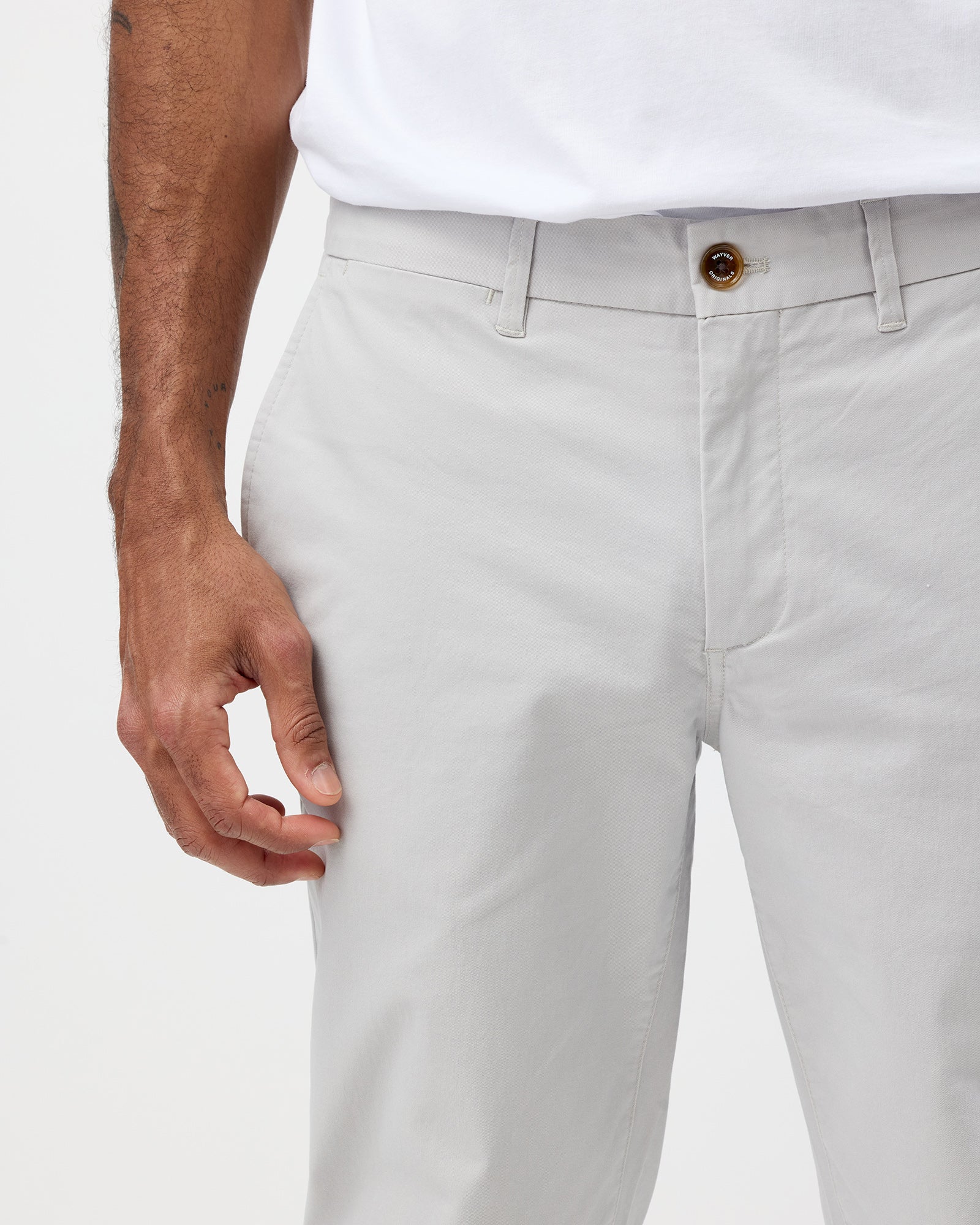 A close-up of a man's hand on his slim stretch chino trouser pocket, wearing a white shirt and gray trousers.
