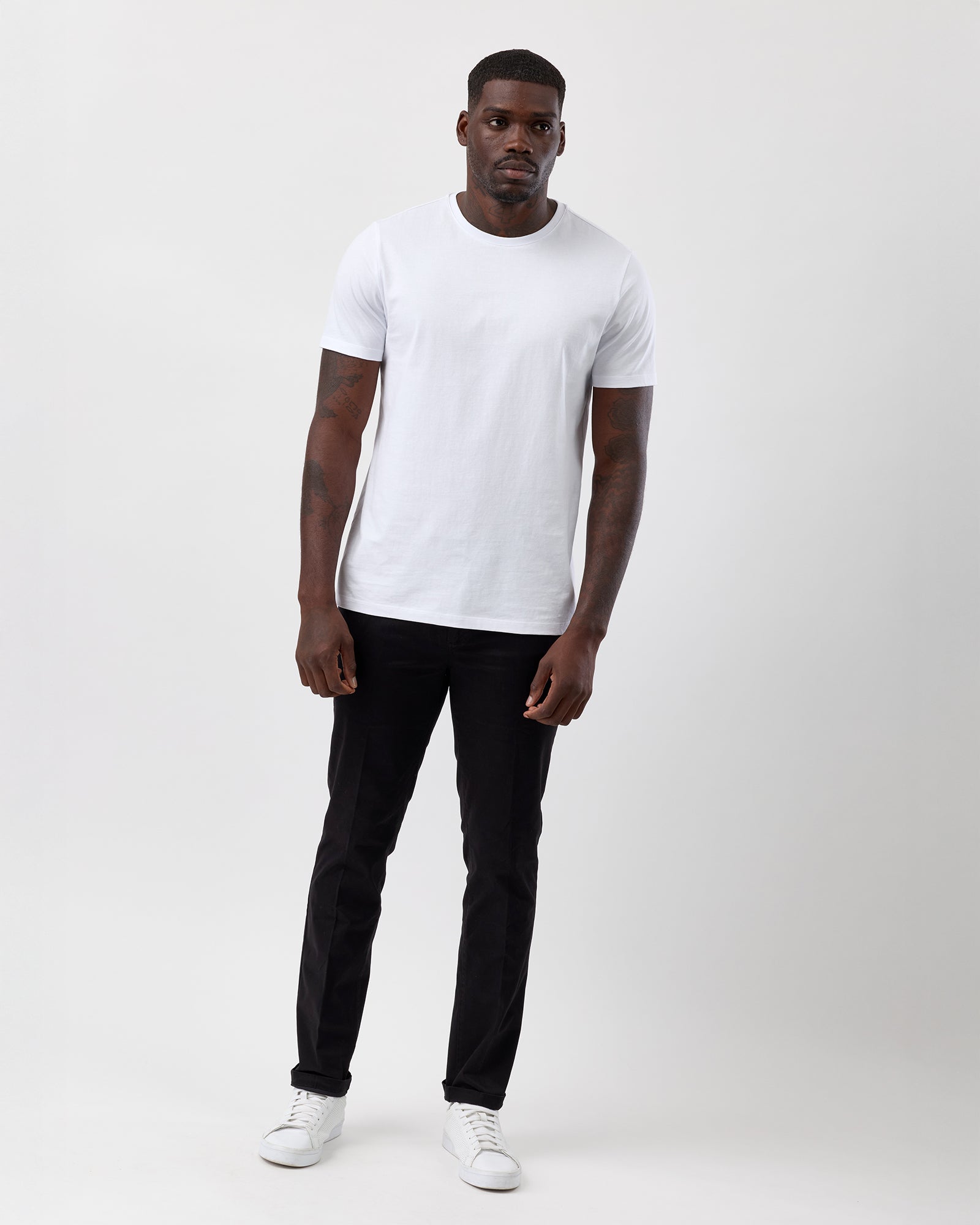 Man wearing a white t-shirt, black cotton stretch chino pants from Wayver, and white sneakers on a plain background, street styled, full body shot