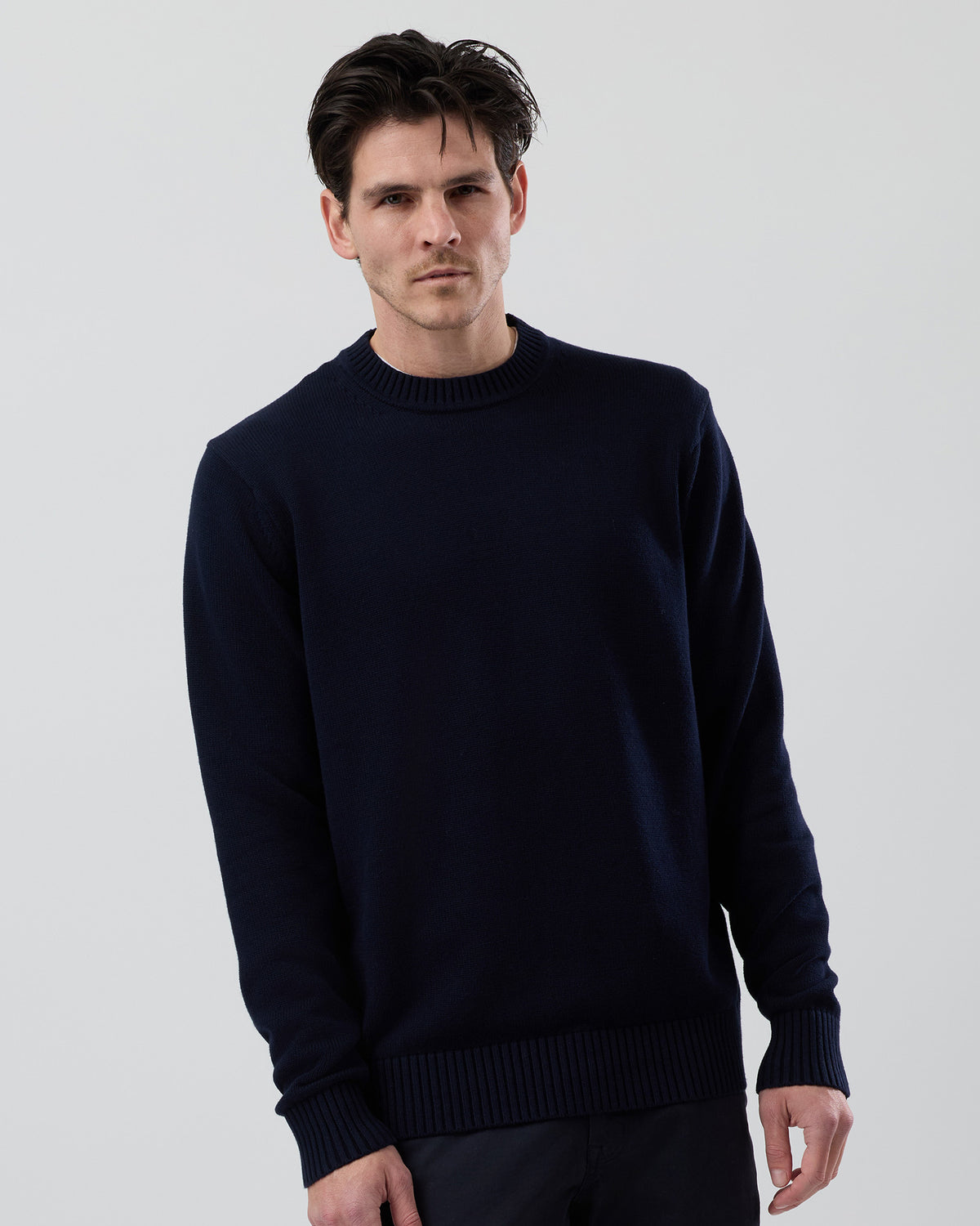 Male model wearing the Wayver Essential Cotton Crew Neck Knit in navy, front view showing the regular fit, slim sleeves and classic crew neck design on a grey background.