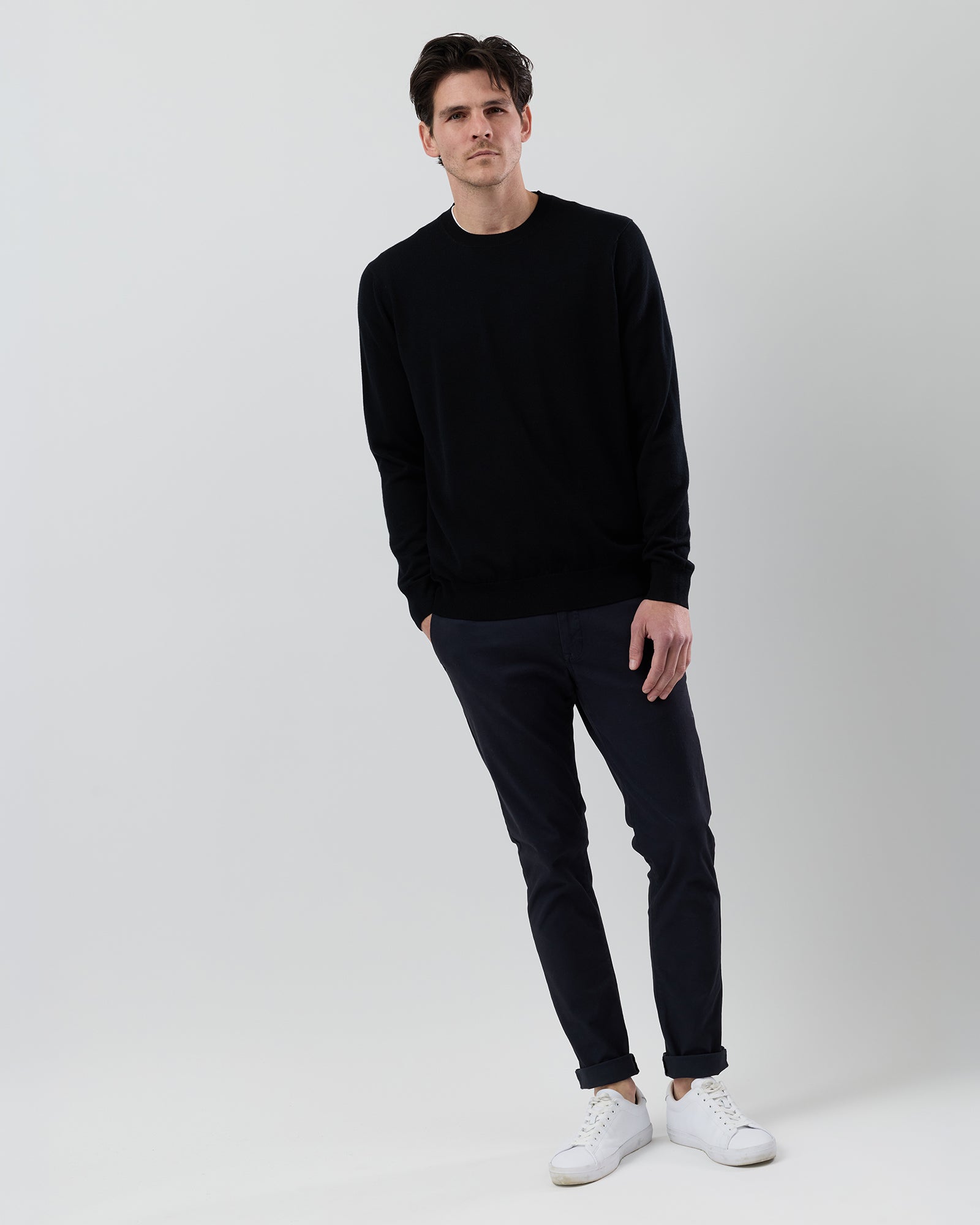 Full-body front view of a male model wearing a black Australian ultrafine merino crew neck knit with navy chinos against a grey background.