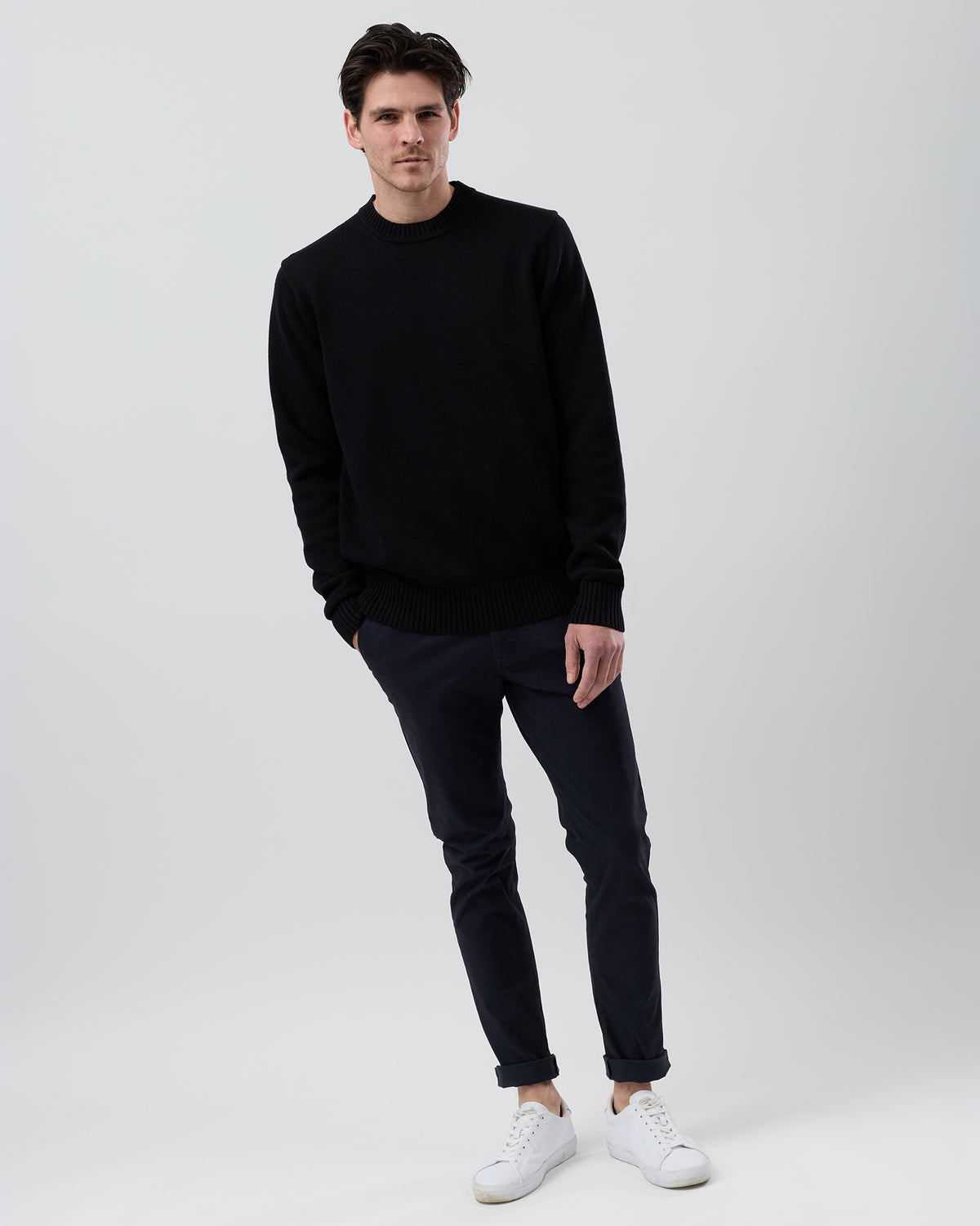 Full-length front view of a male model wearing the Wayver black cotton crew neck knit styled with navy chinos on a grey background.