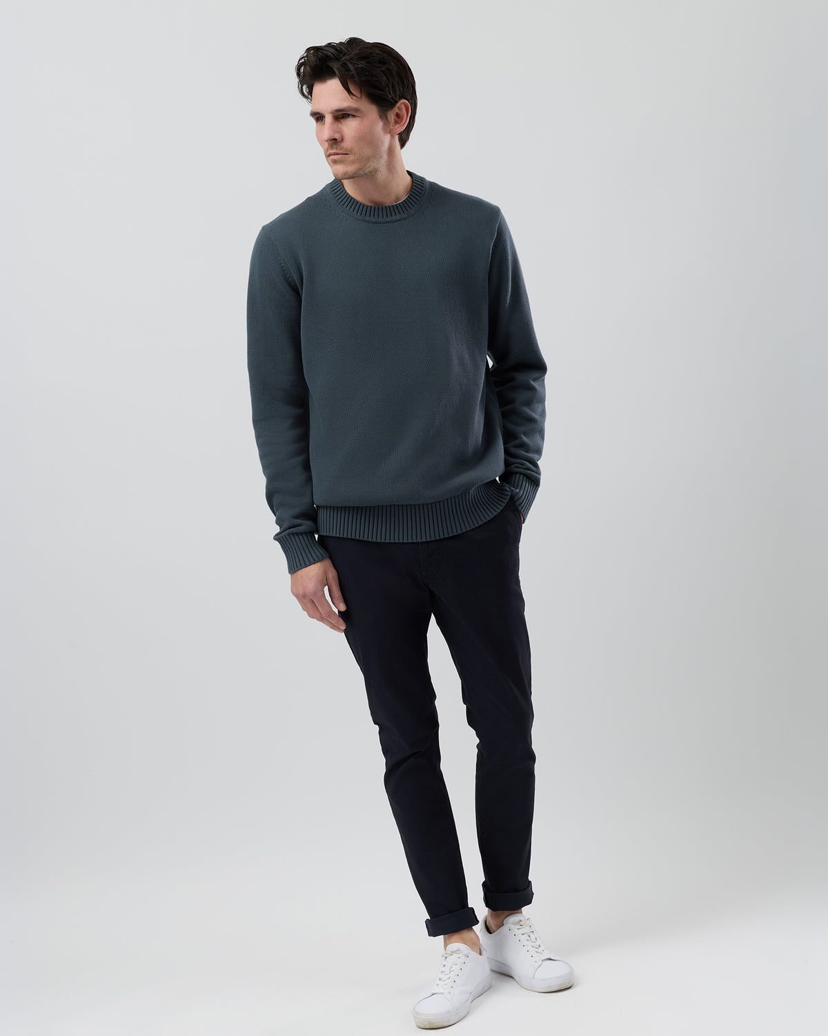 Full body view of a male model wearing the Wayver ironstone Essential Cotton Crew Neck Knit styled with navy chinos on a grey background.