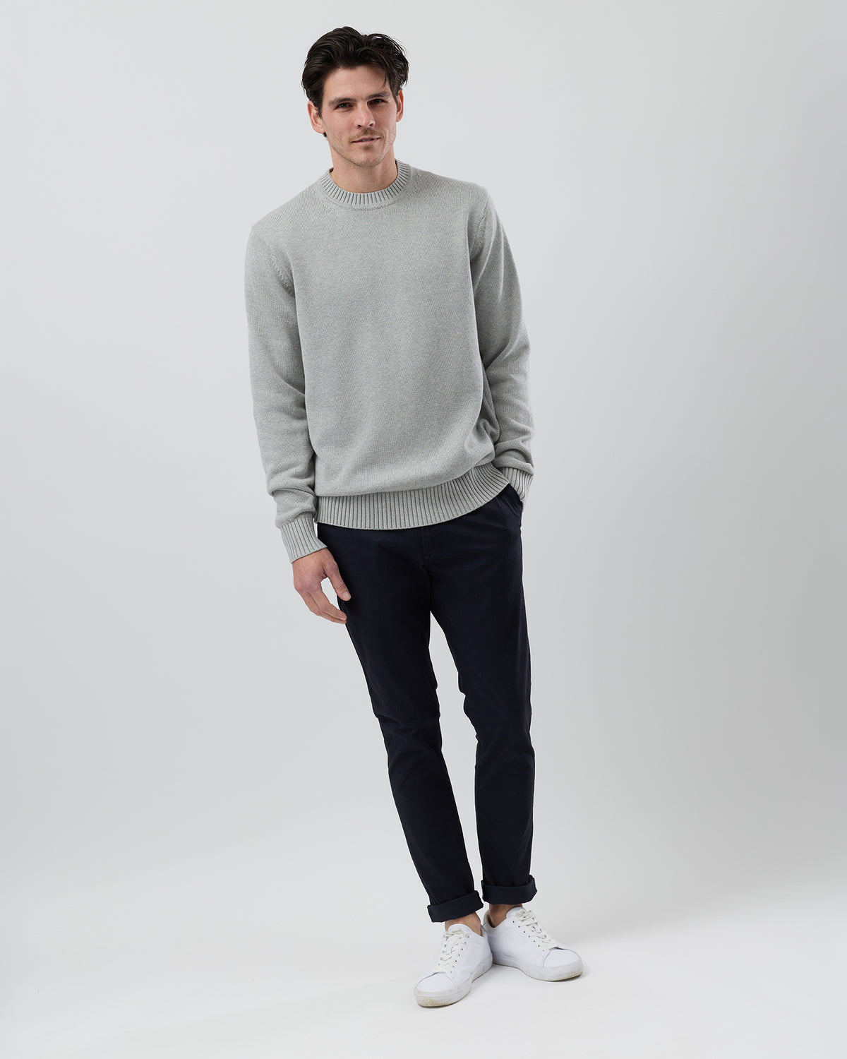 Full body view of a male model wearing the Wayver light grey Essential Cotton Crew Neck Knit styled with navy chinos on a grey background.