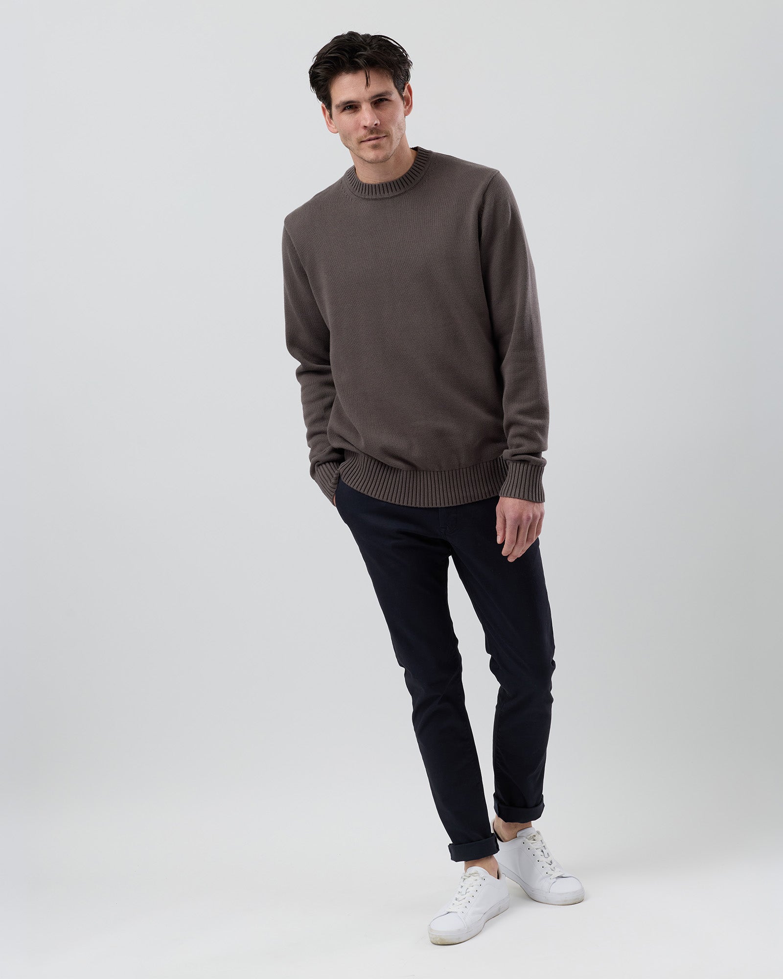 Full body view of a male model wearing the Wayver slate brown Essential Cotton Crew Neck Knit styled with navy chinos on a grey background.