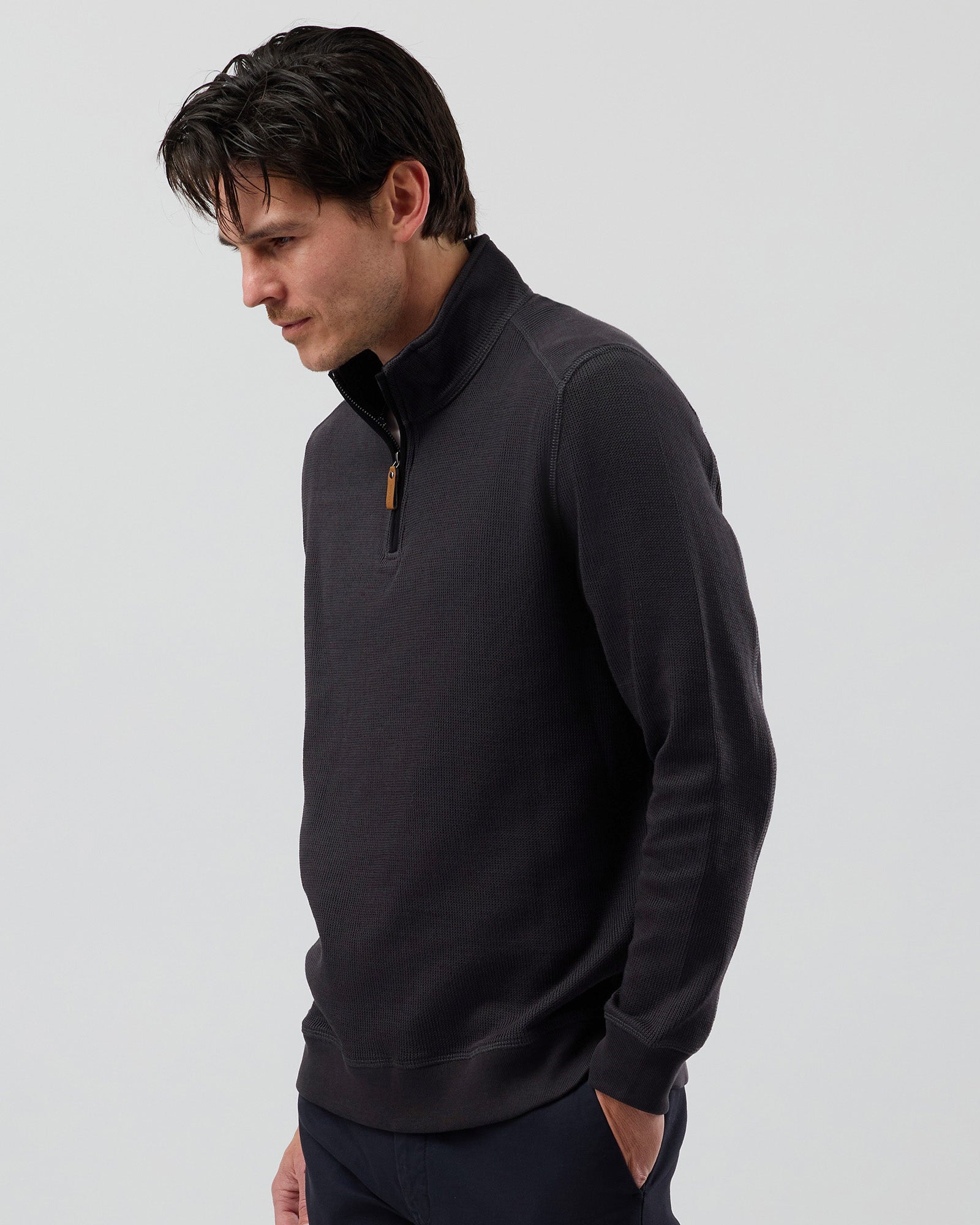 A man wearing a charcoal long-sleeve quarter-zip pullover sweater with a small tan zipper pull, layered over a white undershirt, set against a plain light grey studio background.