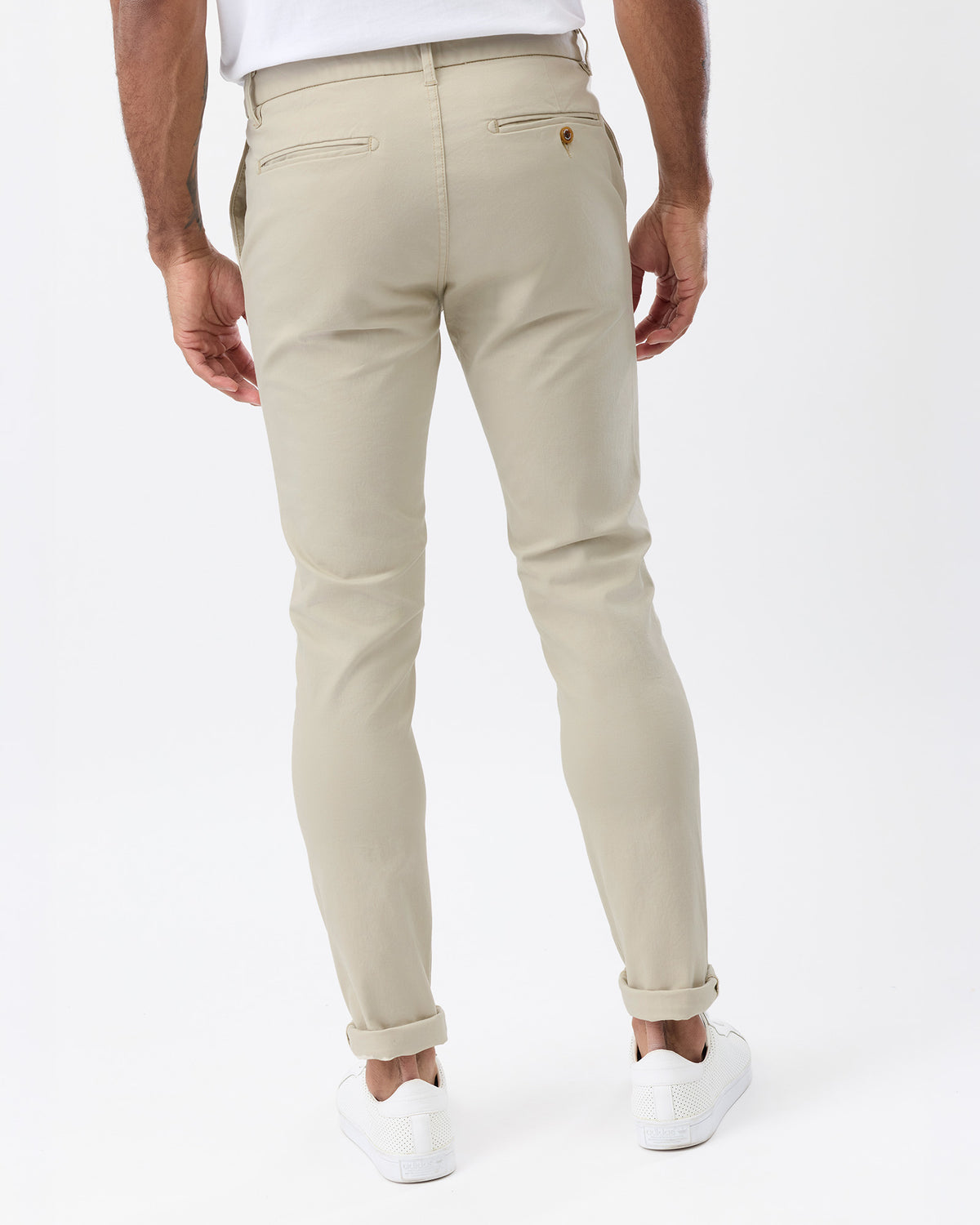 A man wearing beige slim-fit chino pants, shown from the back. The pants feature a classic five-pocket design, belt loops, and rolled cuffs, paired with a white t-shirt and white sneakers against a clean white studio background.
