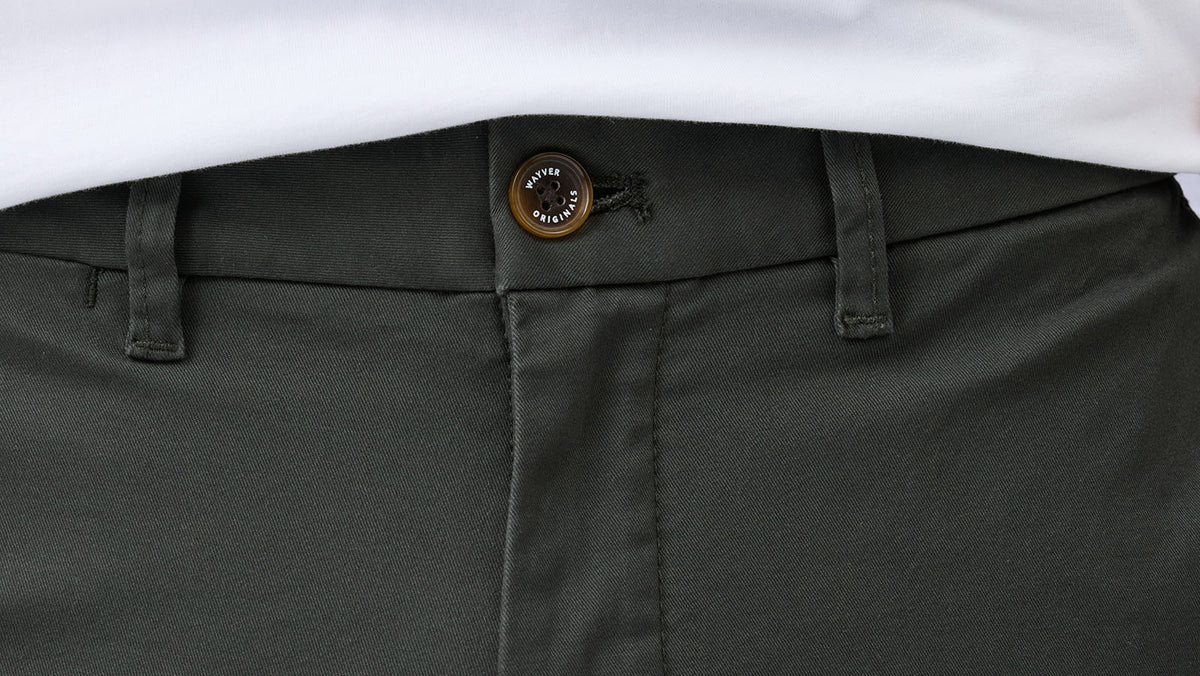 Close-up of Wayver Originals vintage green cotton stretch chinos showing the waistband, branded button, belt loops and twill fabric.
