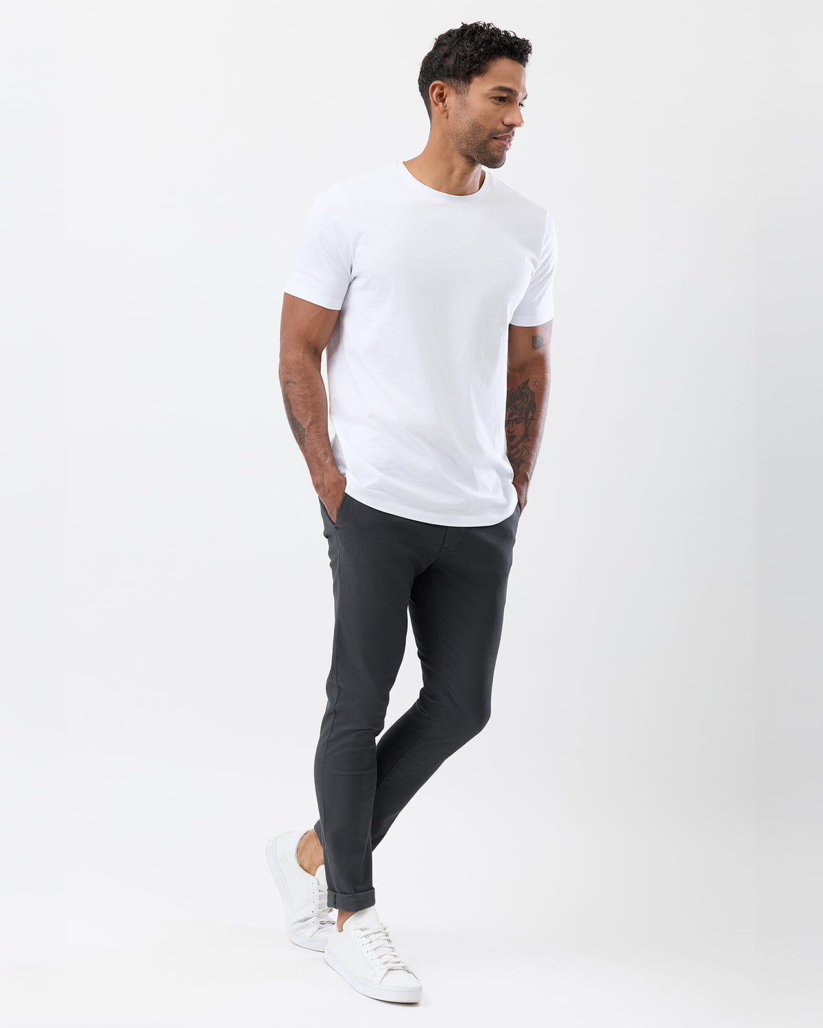 A man wearing a white crew-neck t-shirt, dark charcoal gray slim-fit pants, and white sneakers, standing in profile against a clean white studio background.