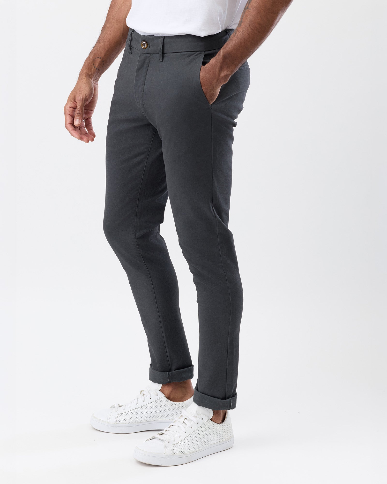 A person wearing slim-fit dark grey (charcoal) trousers, paired with a white t-shirt and white sneakers, shown from the waist down on a light grey studio background.